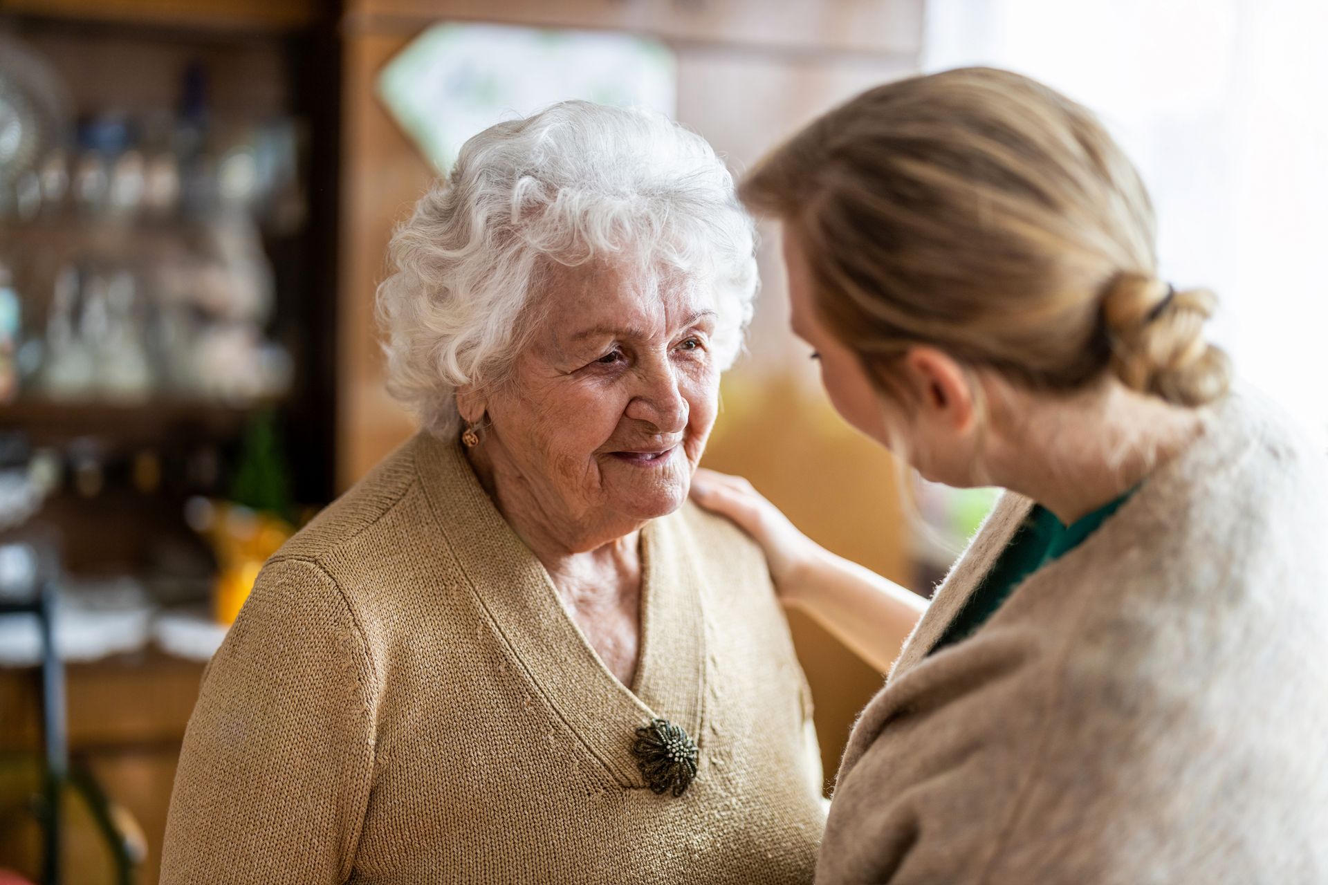 A woman warmly comforts an elderly woman in a cozy indoor setting.