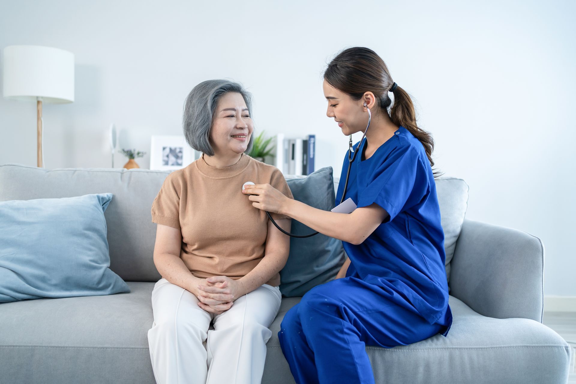 Nurse in blue scrubs checks an older patient's chest on a couch in a bright living room.