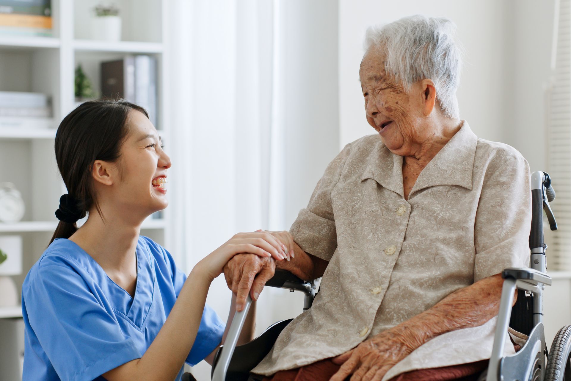 Caregiver in blue scrubs smiling and holding hands with an elderly woman in a wheelchair in a bright room