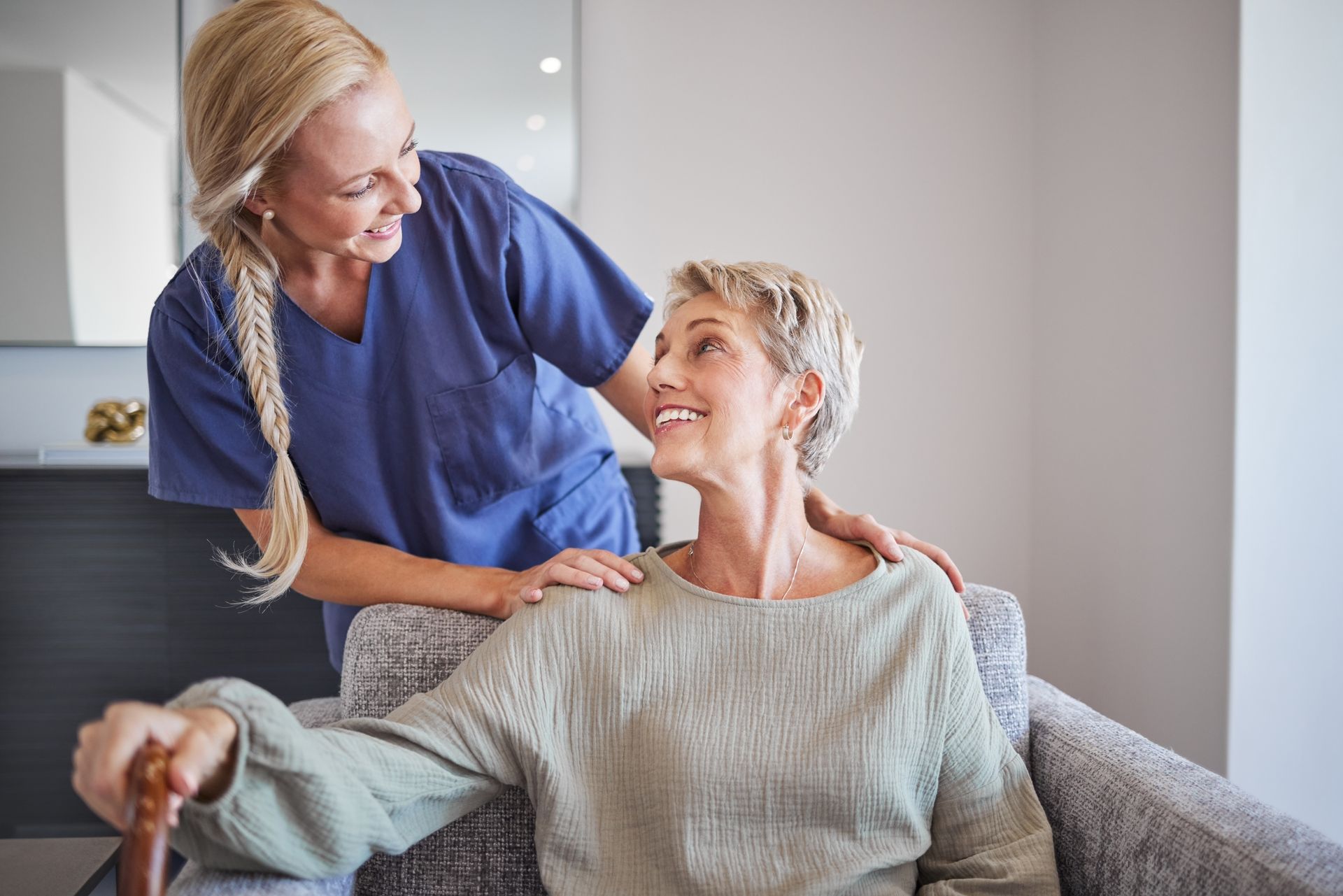 Caregiver in blue smiling and adjusting a blanket around a seated woman in a cozy living room