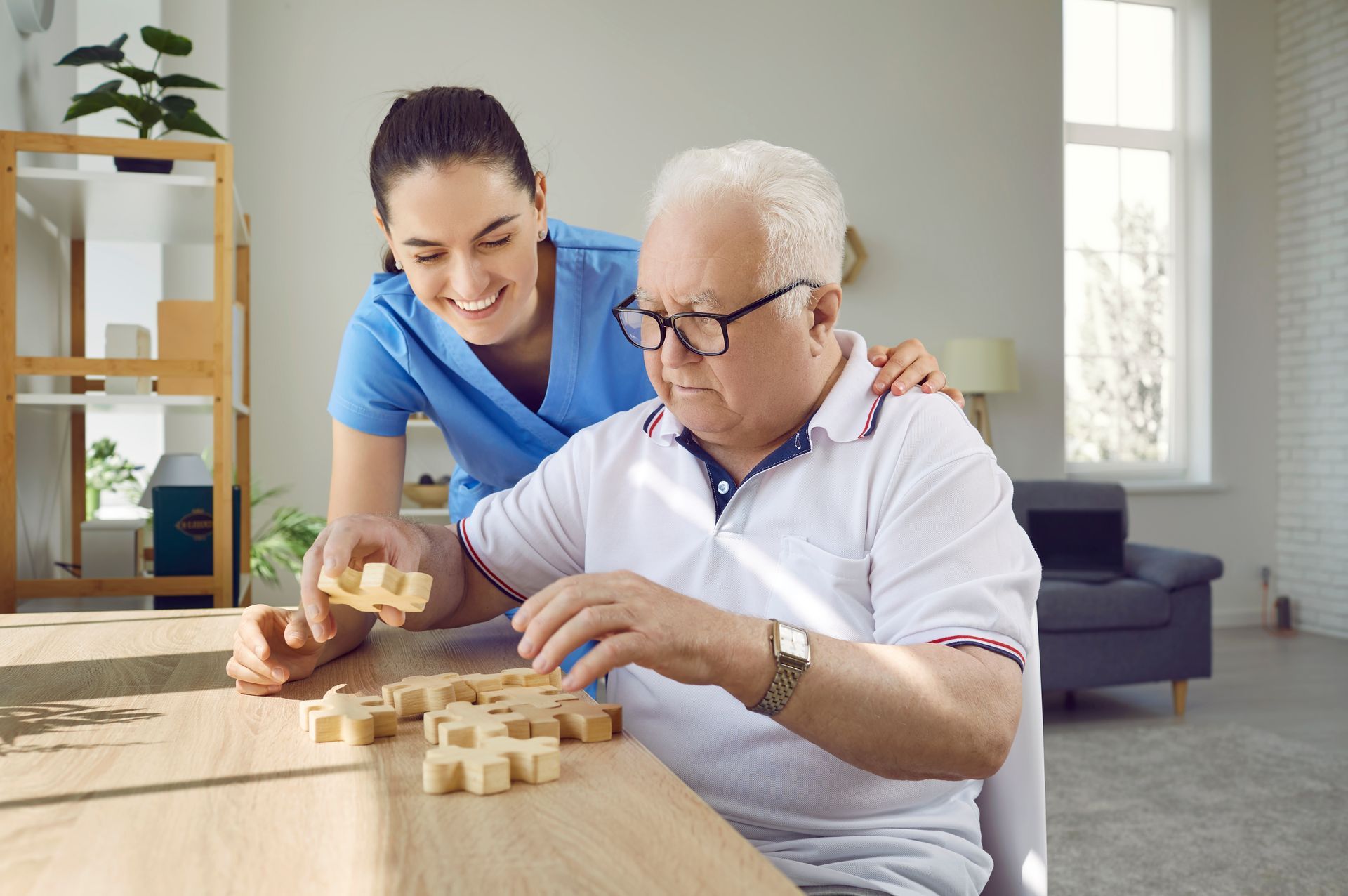 Caregiver helping an older man play a wooden block game at a table in a bright living room