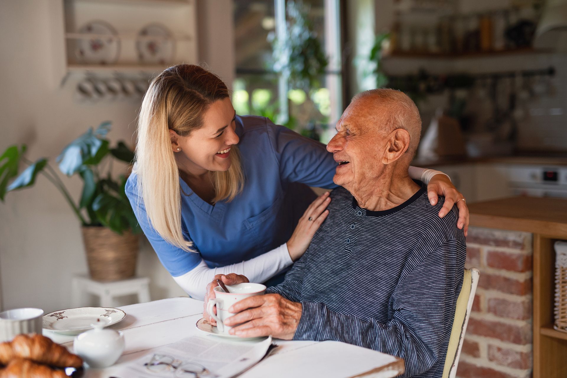 A caregiver smiles, her arm around an elderly man holding a mug at a kitchen table.