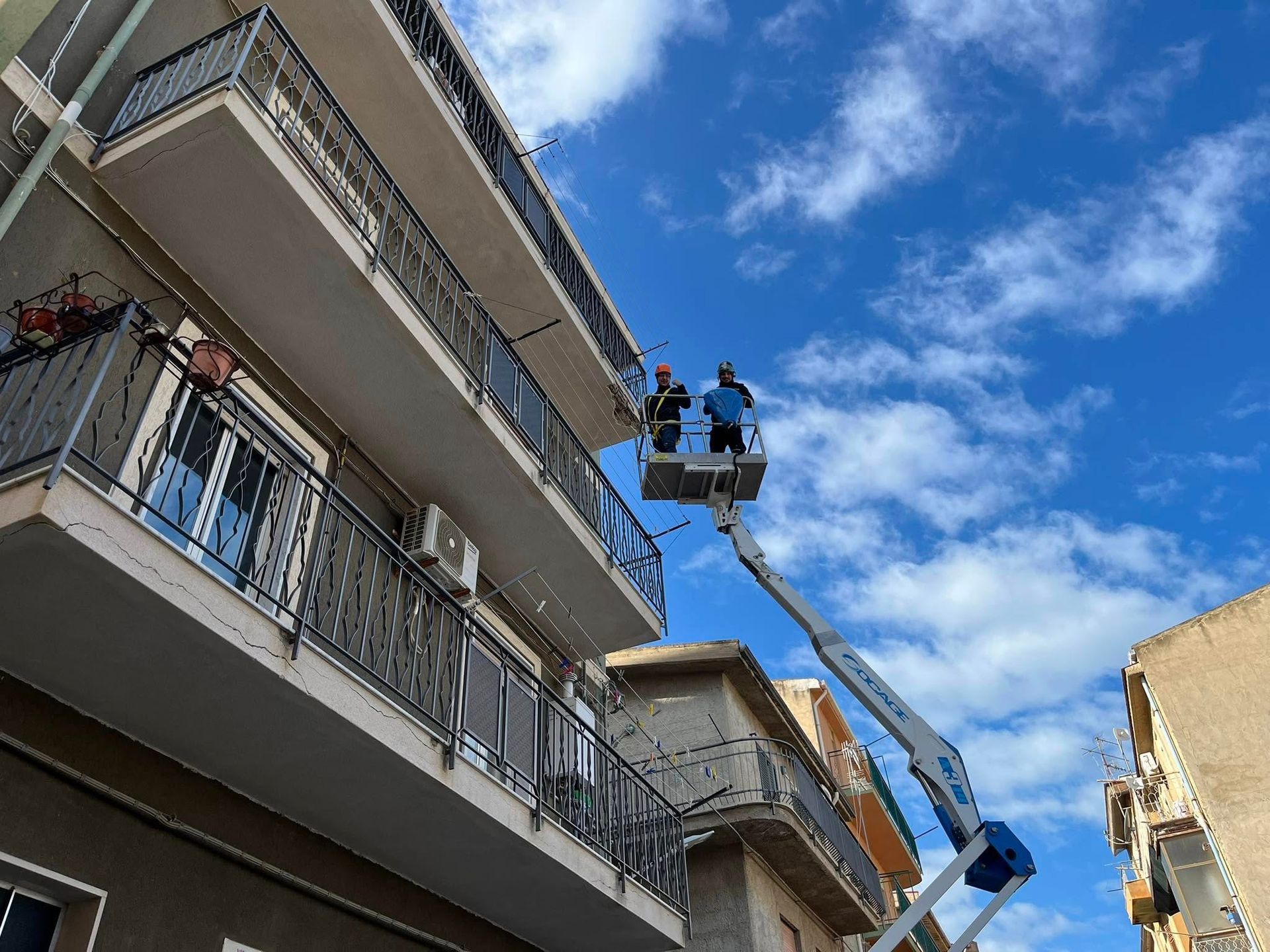 Due persone su una piattaforma elevatrice accanto a un edificio, lavorano sul balcone più alto sotto un cielo azzurro e nuvoloso.