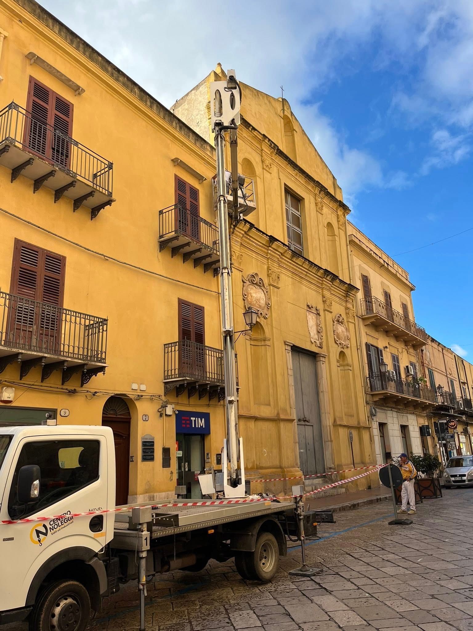 Camion con sollevatore accanto a un edificio giallo. Operai nelle vicinanze. Cielo azzurro brillante.