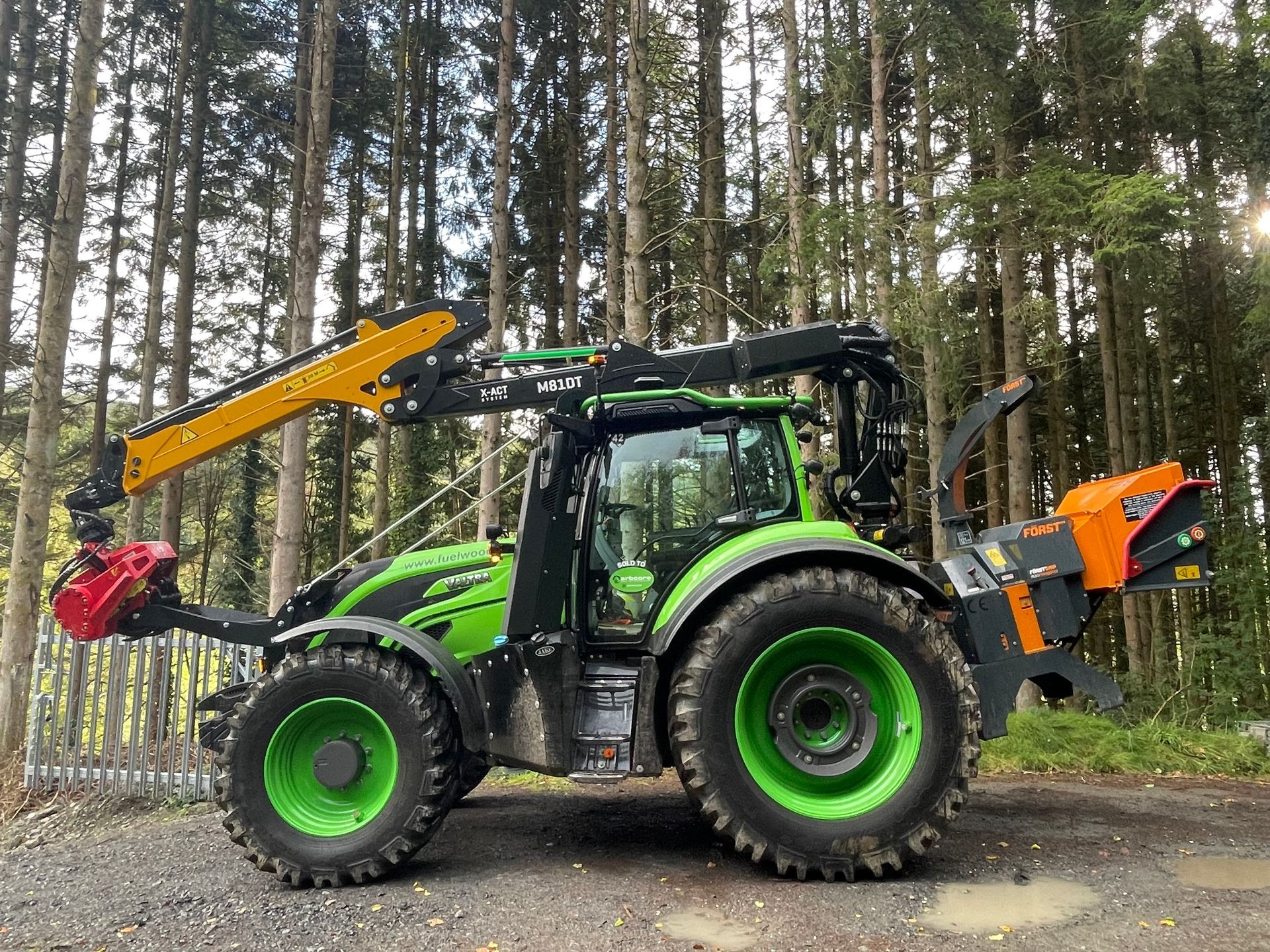 Green tractor with loader and rear attachment parked on a gravel road in a pine forest