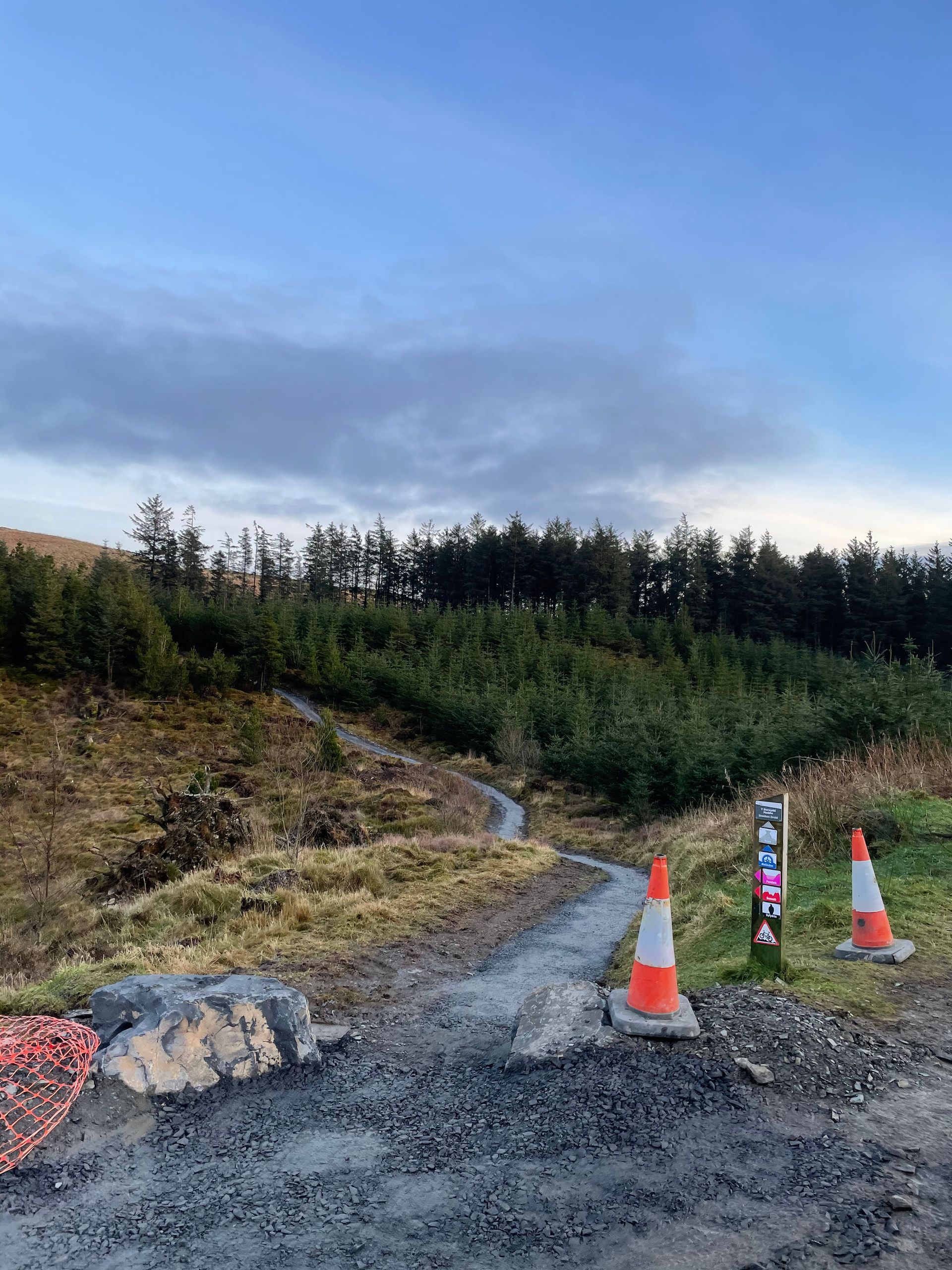 Gravel path in a rural area with orange traffic cones and a forested hillside under a blue sky