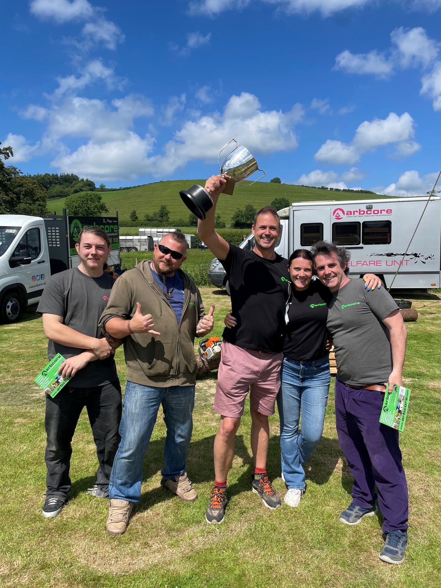 Five people smiling outdoors, holding drinks and a trophy in front of a van under a blue sky.