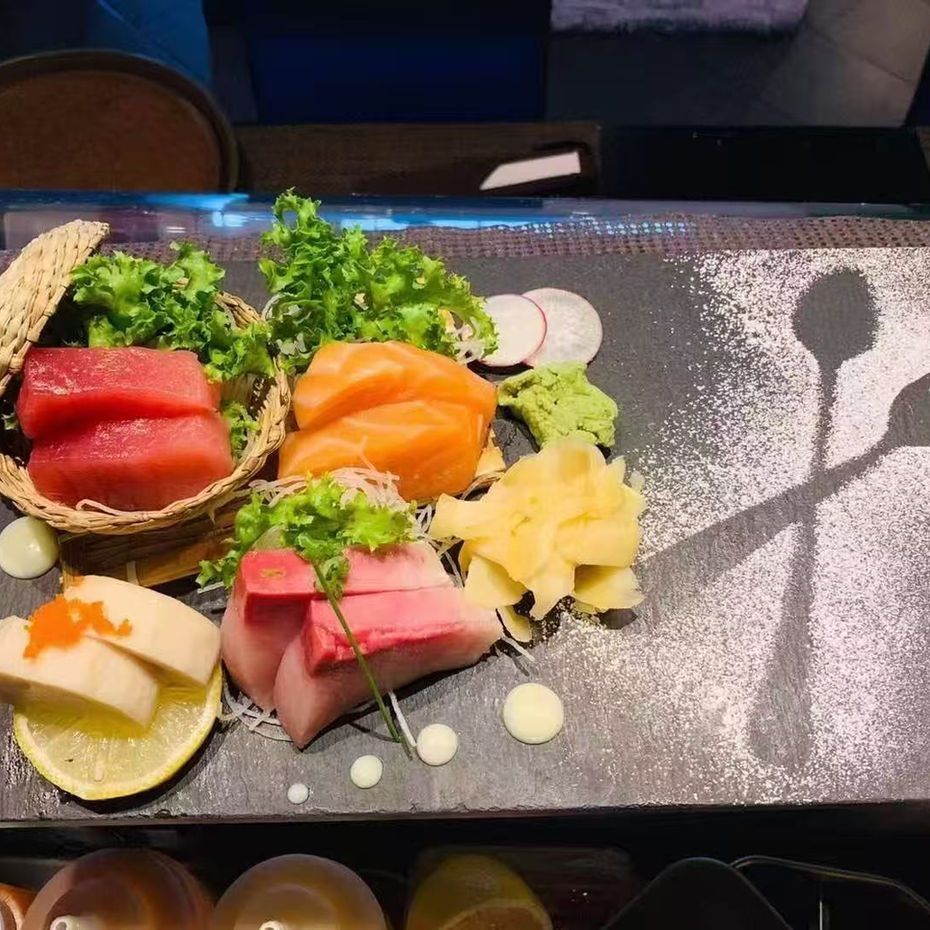 Assorted sashimi and seafood served on a black platter with lemon, garnish, and decorative sauce dots