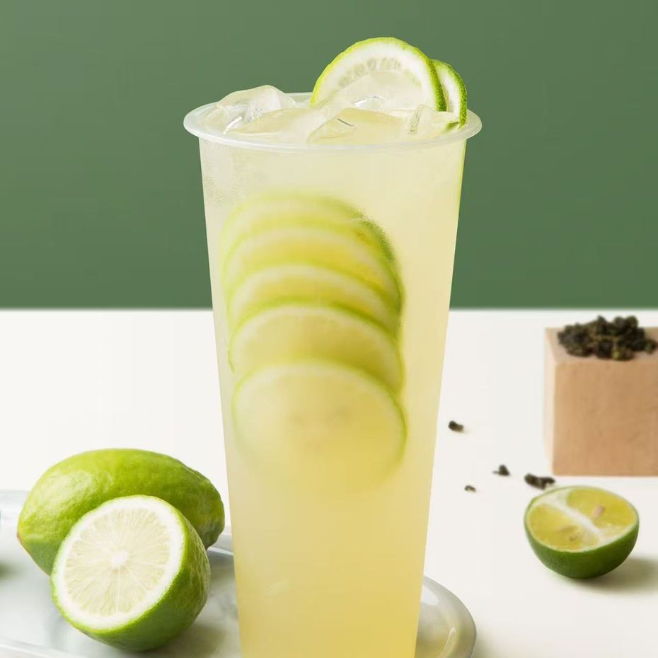 Lime drink in a tall glass with lime slices, ice, and whole limes on a table against a green background