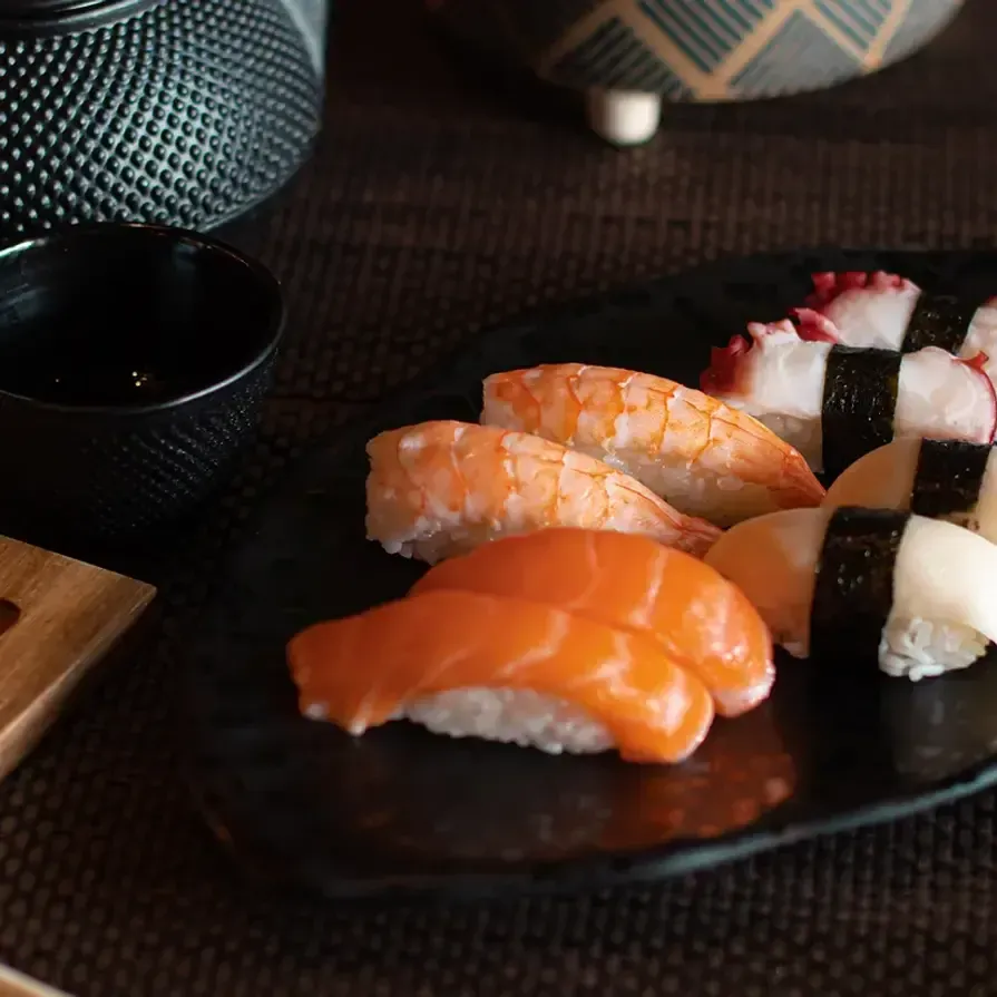 Assorted sushi nigiri on a dark plate with a soy sauce cup on a woven table setting