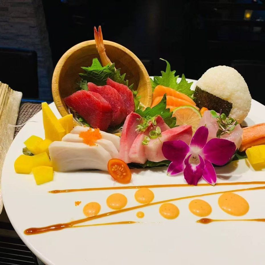 Assorted sushi and sashimi platter with mango, tuna, shrimp, rice, and orchid garnish on a white plate