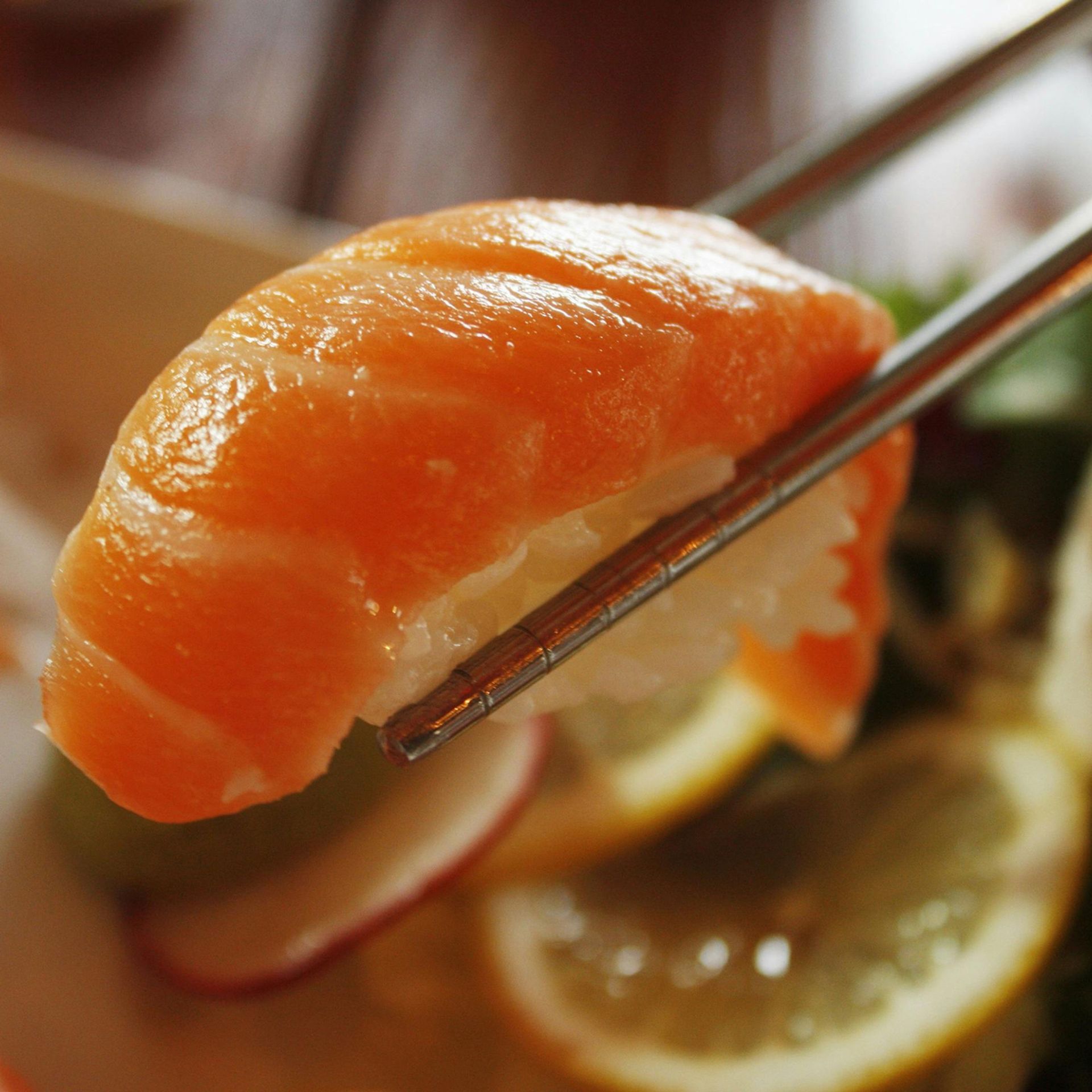 Chopsticks holding a piece of salmon sushi over a lemon slice and sauce dish