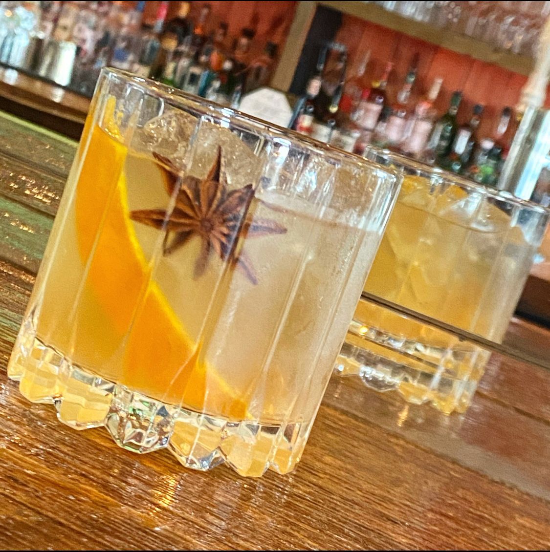 A close up of two drinks on a bar with bottles in the background