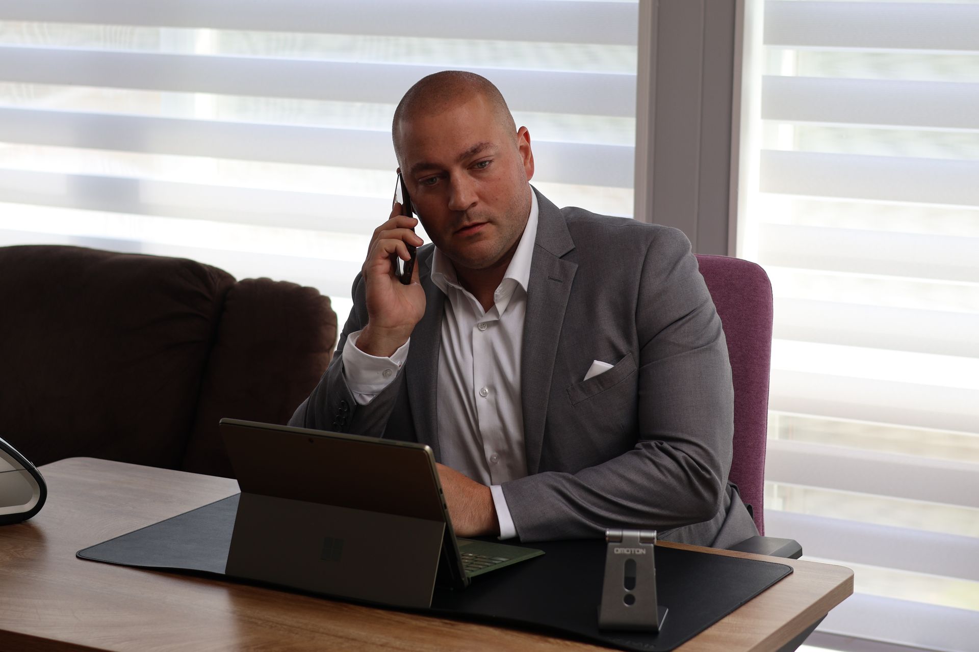 Man in gray blazer talks on phone at a desk with a laptop in a bright office