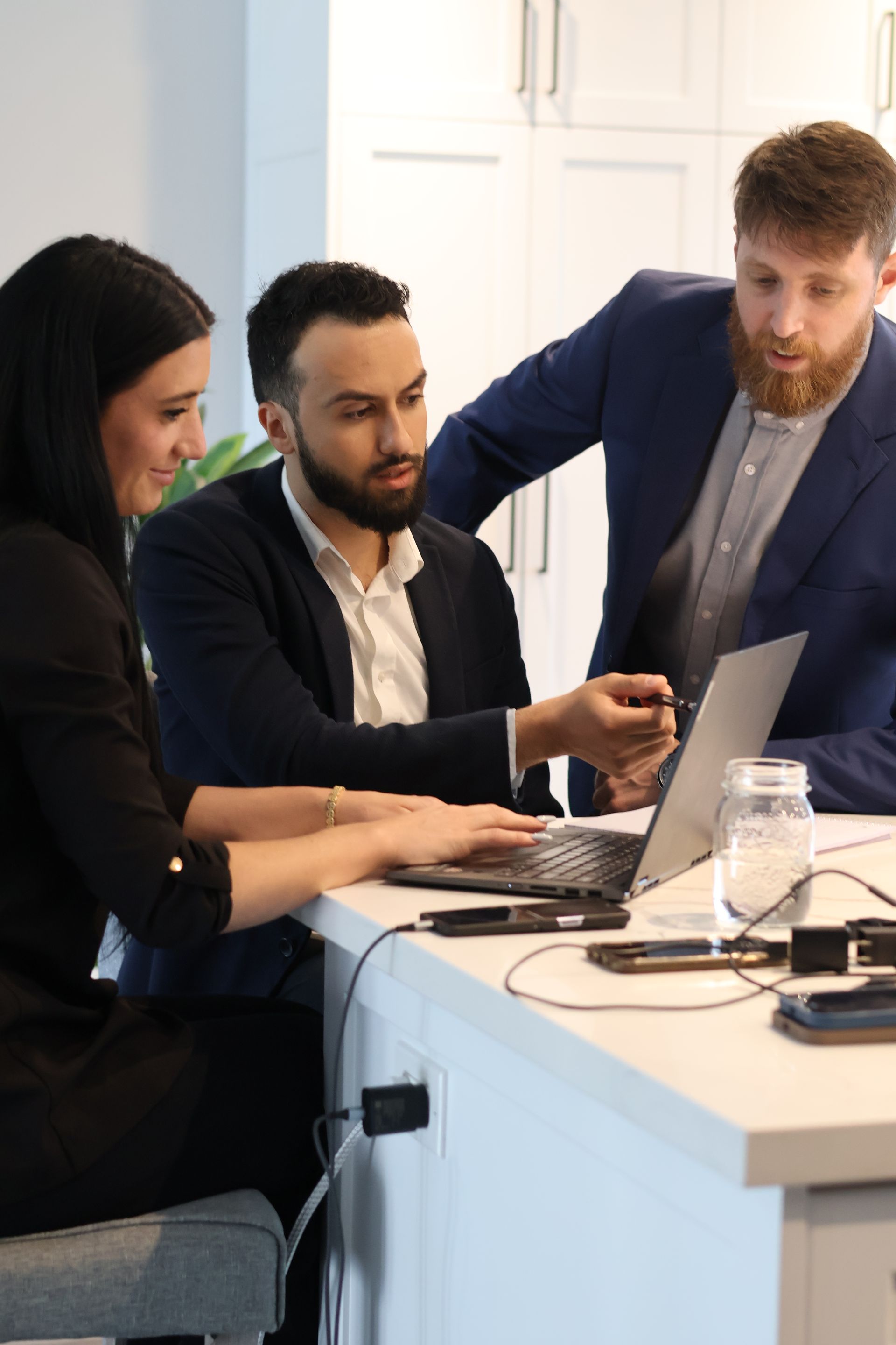 Three coworkers collaborate around a laptop at a kitchen counter in a bright office.