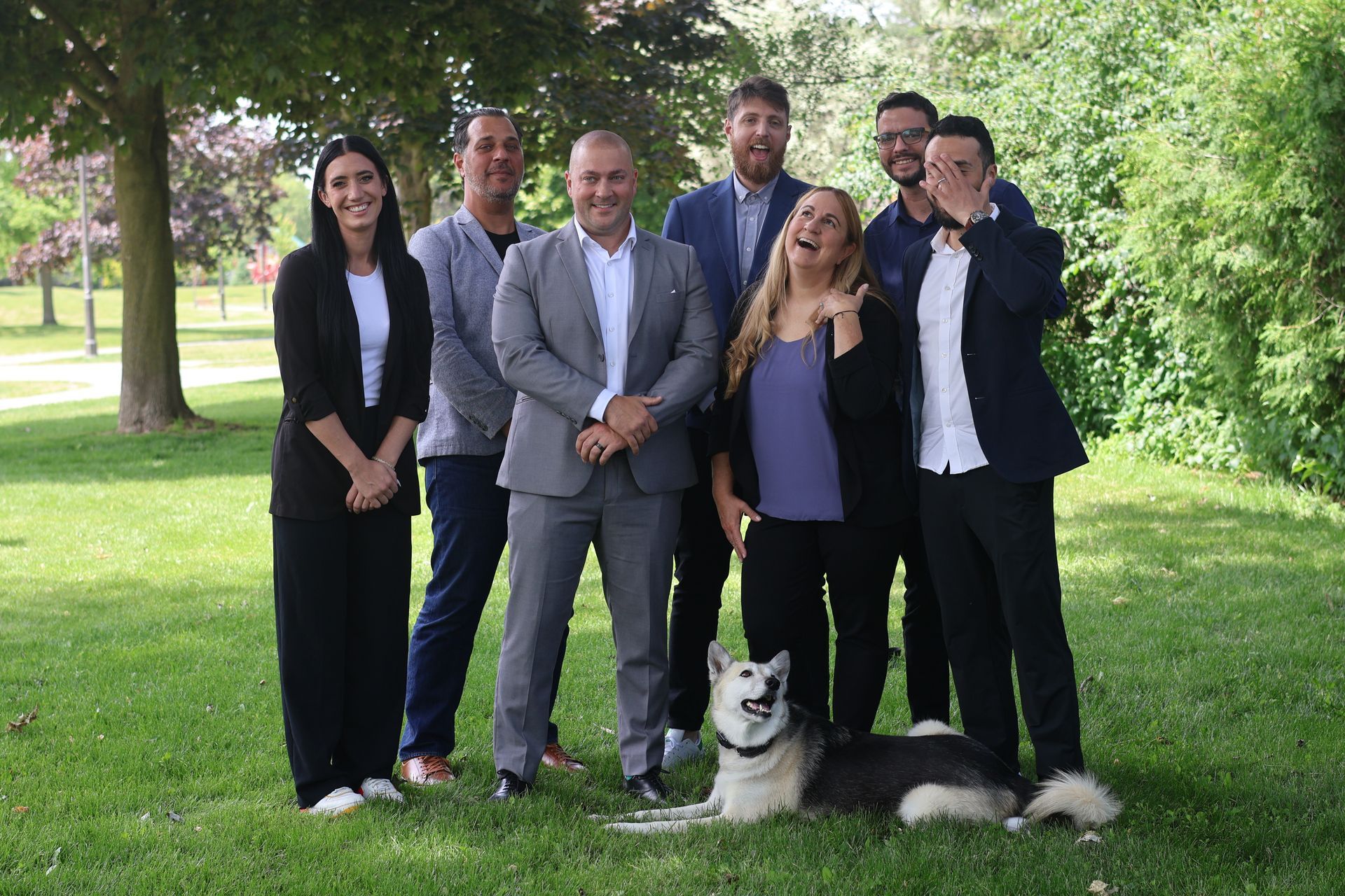 Six people in business attire pose on a grassy lawn with a black-and-white dog in the foreground.