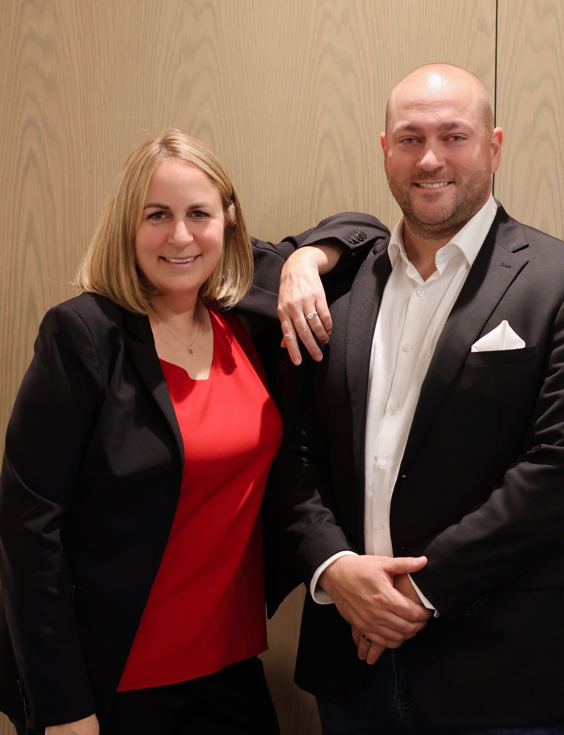 Two people smiling indoors, dressed in black and red business attire against a wood-paneled wall.