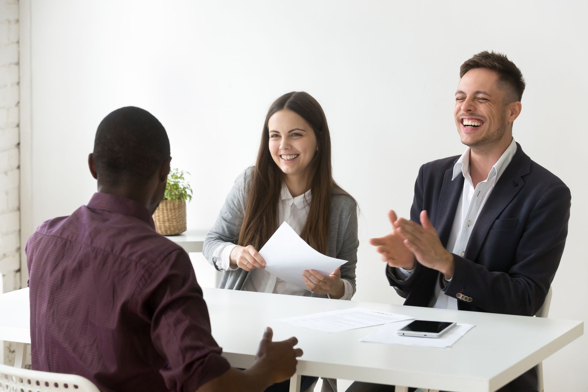Three people in a meeting room discussing papers at a white table, one smiling and clapping.