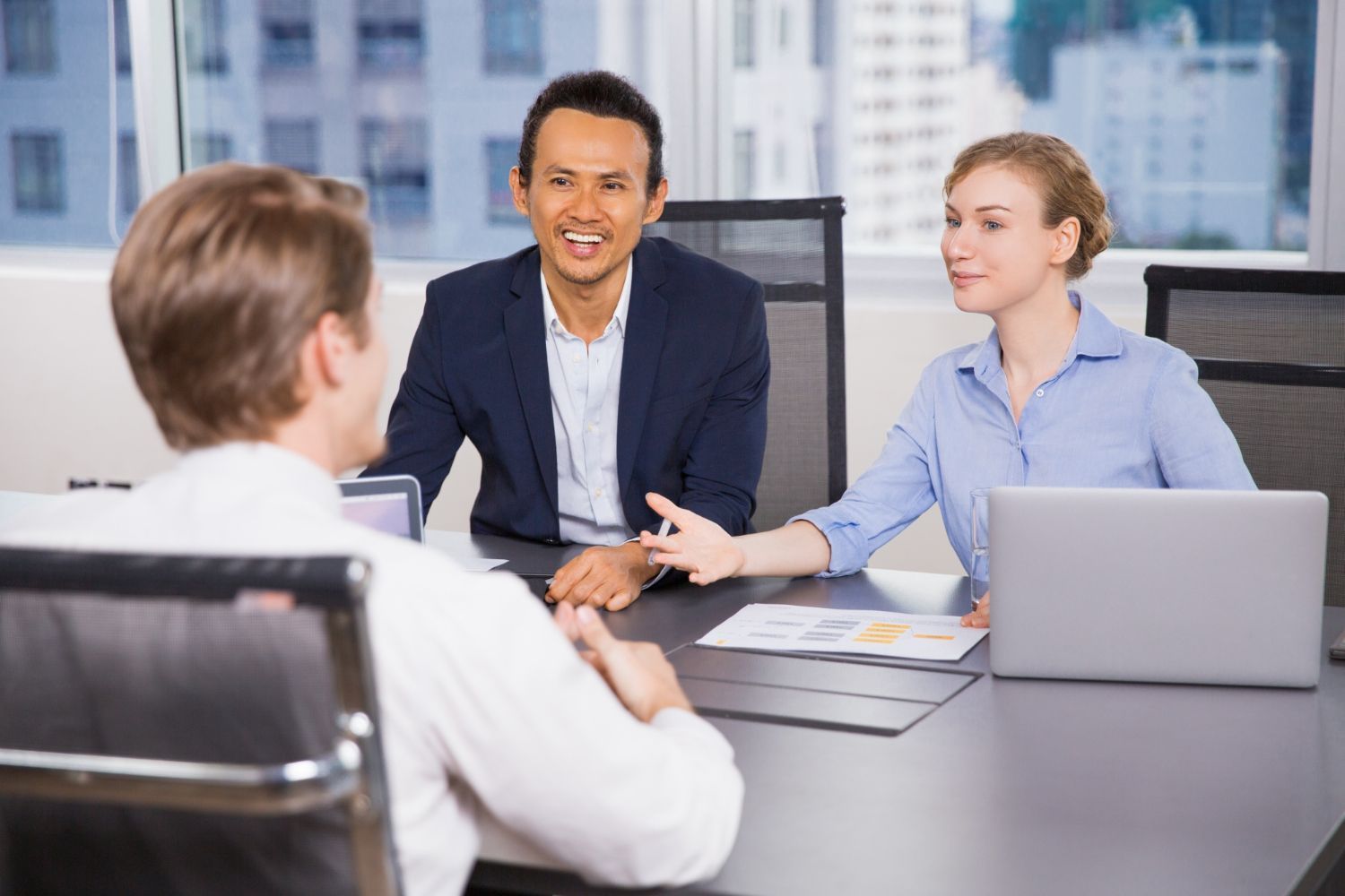 Three coworkers in a conference room discussing a project around a table with a laptop and documents