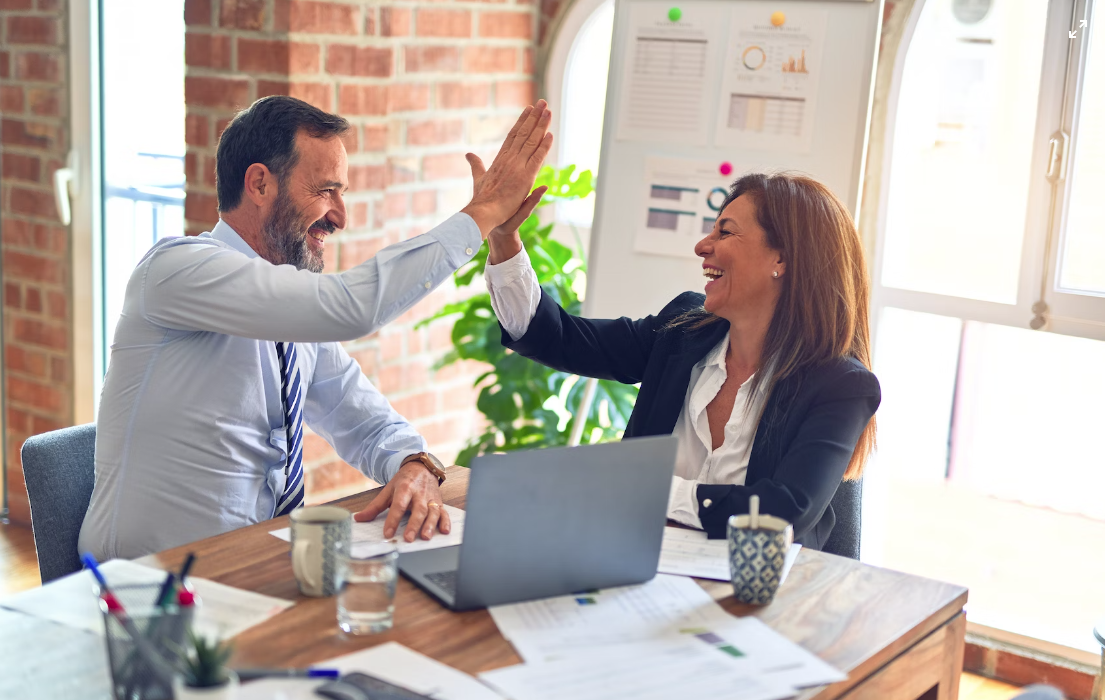 Two coworkers high-five across a desk in a bright office with a laptop and coffee mugs.