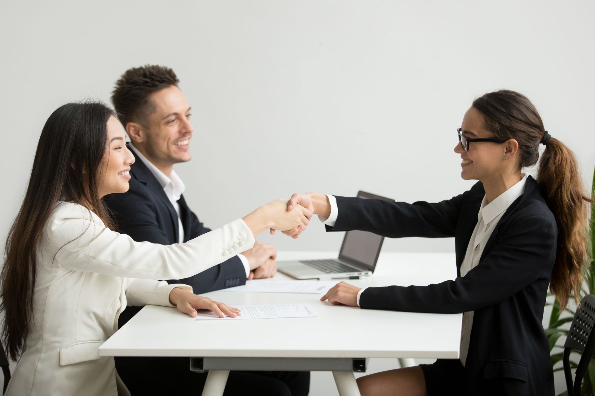 Three people in business attire shaking hands across a meeting table in a bright office