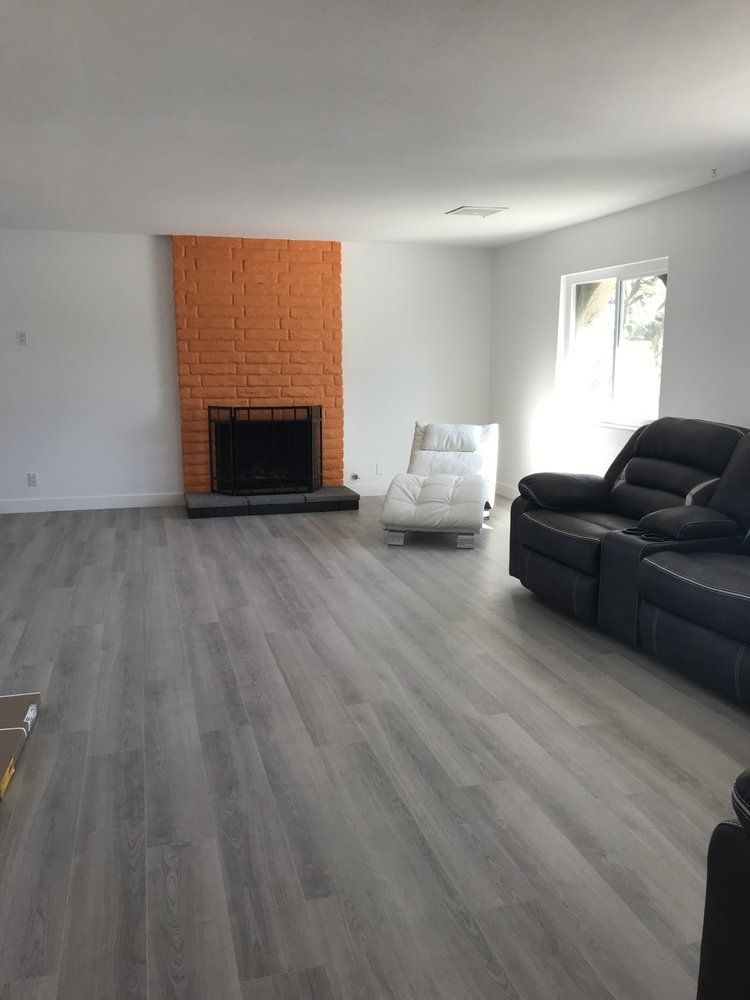 An empty living room with gray flooring, white walls, and an orange brick fireplace. A black leather sofa is on the right.