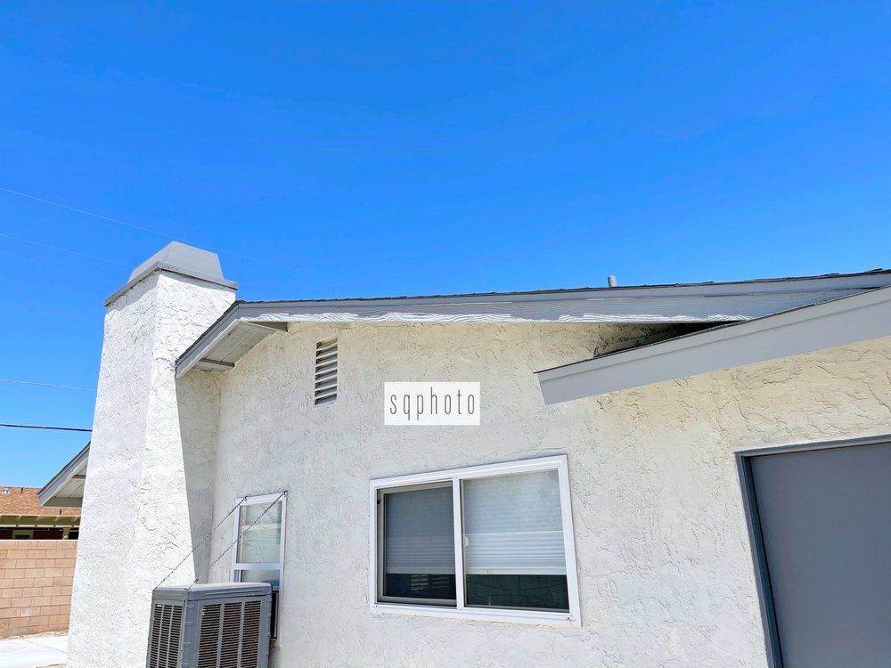 Exterior view of a stucco building with a chimney, window, and air conditioning unit against a blue sky.