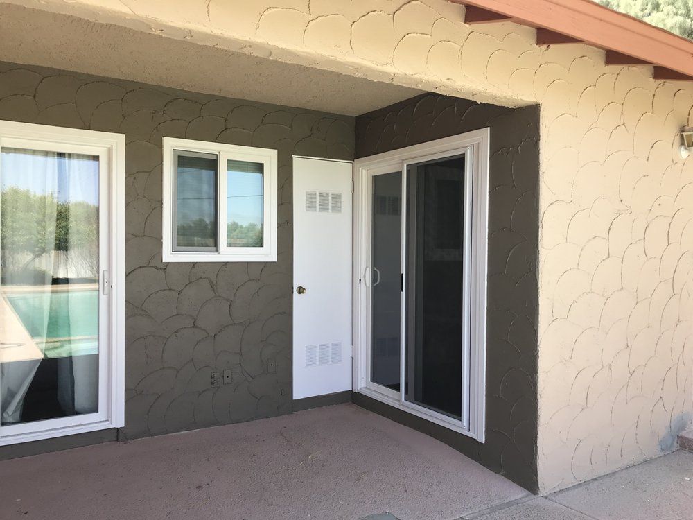 Exterior of a building with white-framed windows and a sliding door. The walls are textured with brown and tan paint.