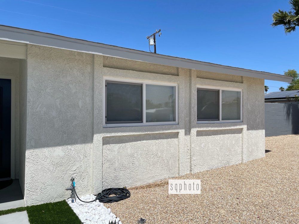 Exterior view of a stucco house with two windows and gravel yard under a blue sky.