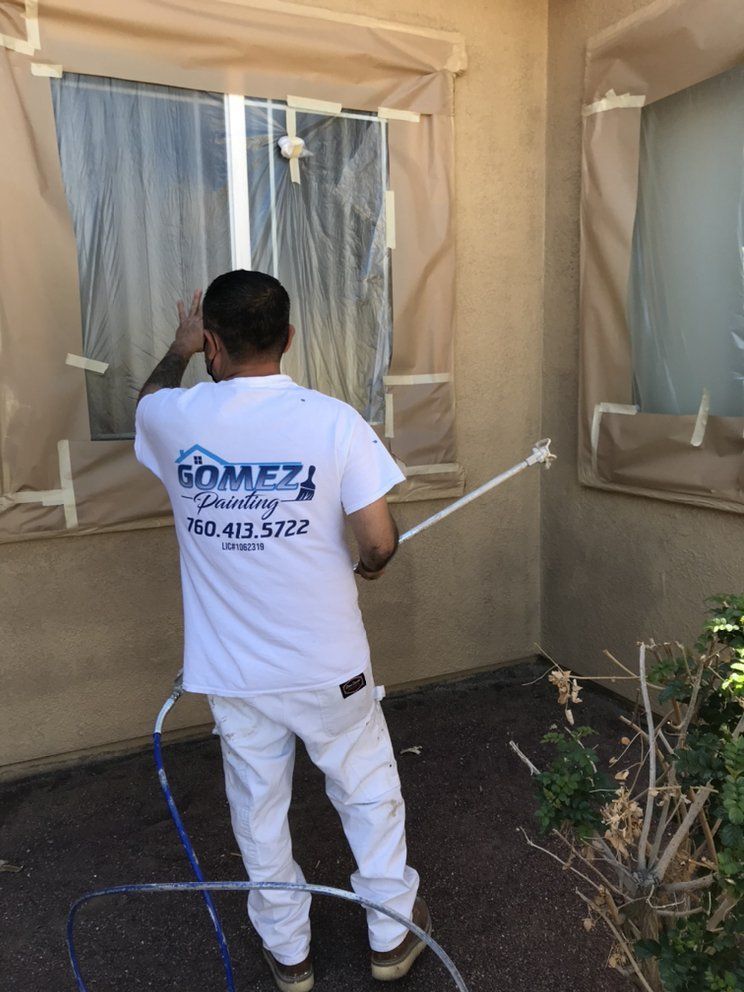 Man painting exterior wall with spray gun; windows taped off, tan wall.