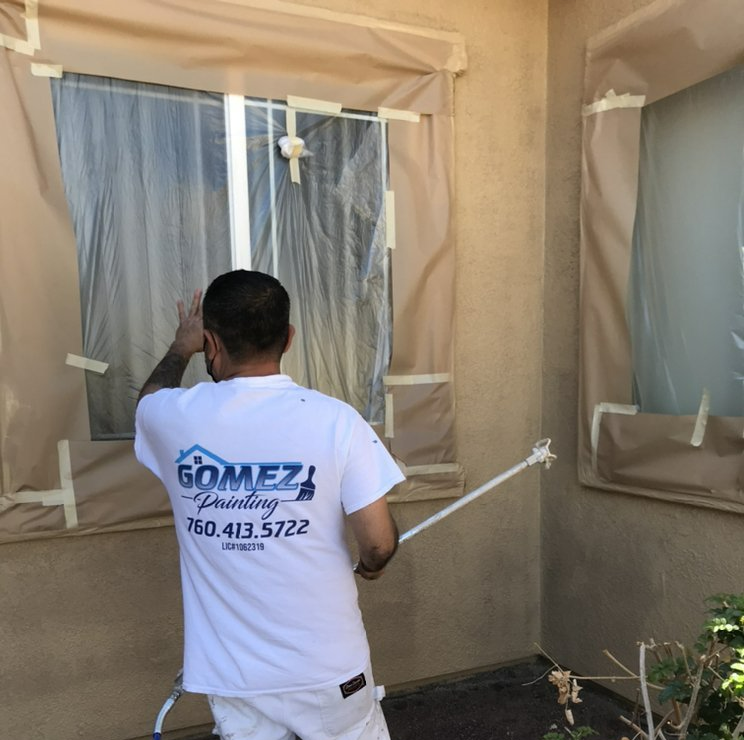 Person spraying exterior wall near windows. Brown tape, tarp, white trim, tan wall.