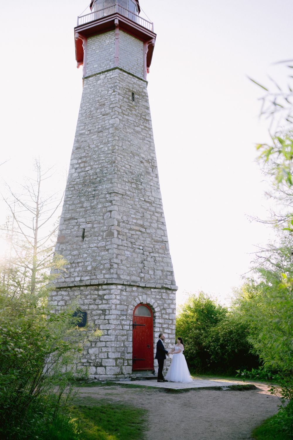 A bride and groom are standing in front of a lighthouse.