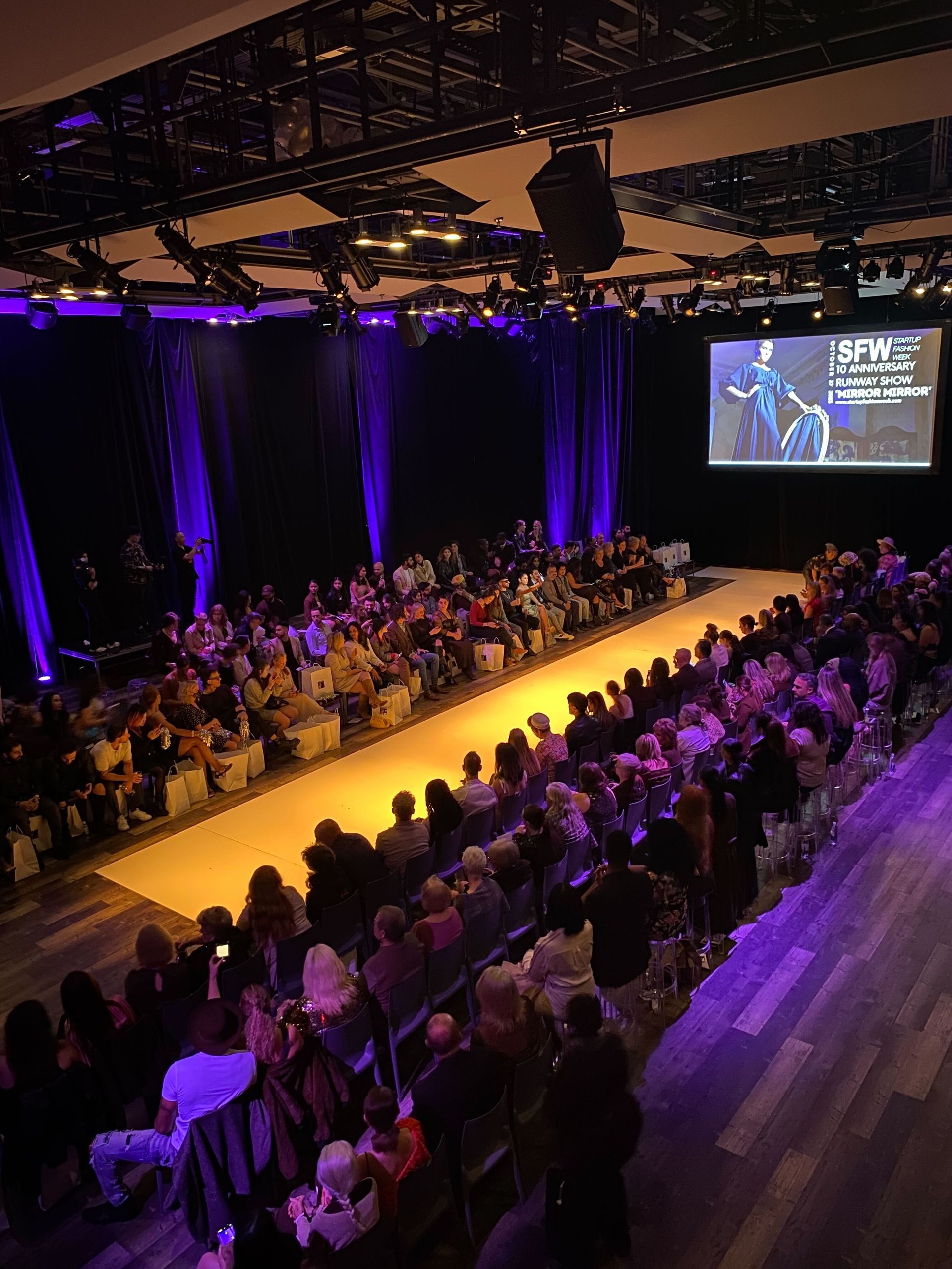 A crowd of people are sitting in a large auditorium watching a fashion show.