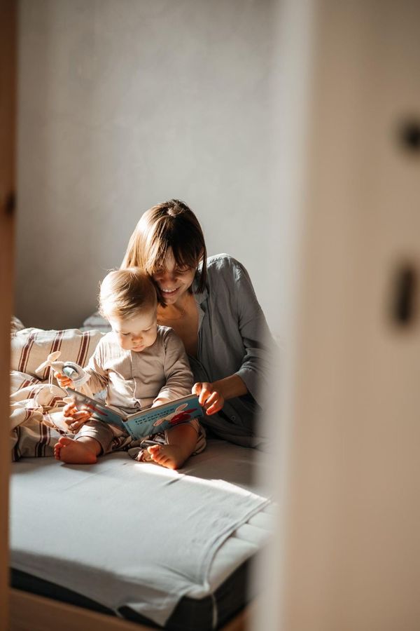 Adult and child sitting on a bed, reading a book in warm sunlight