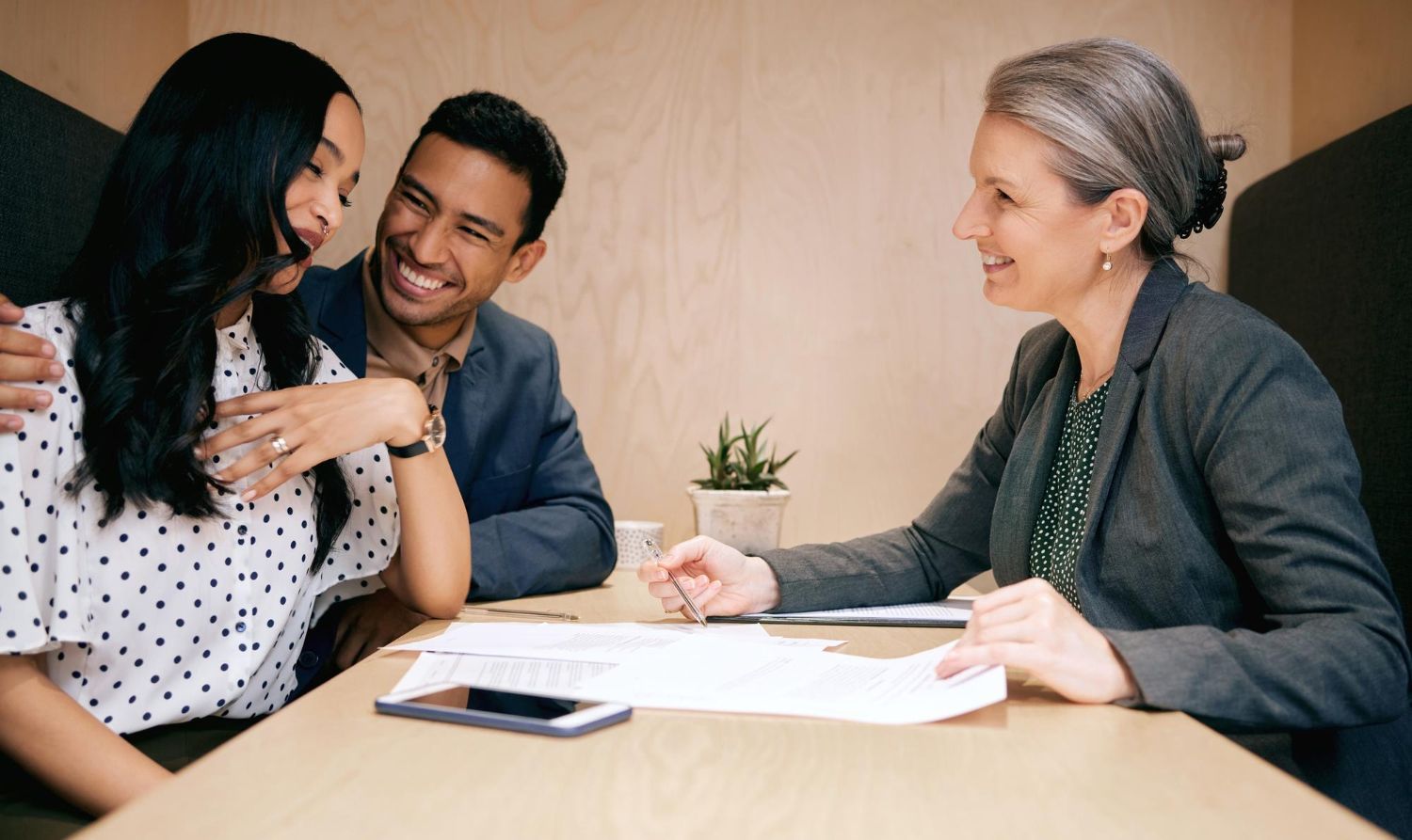 Three people in a meeting, smiling and reviewing documents at a table.