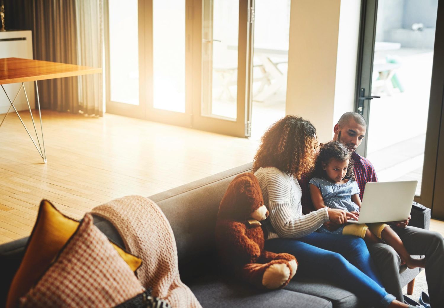 Family sitting on a couch using a laptop in a sunlit living room with a teddy bear nearby