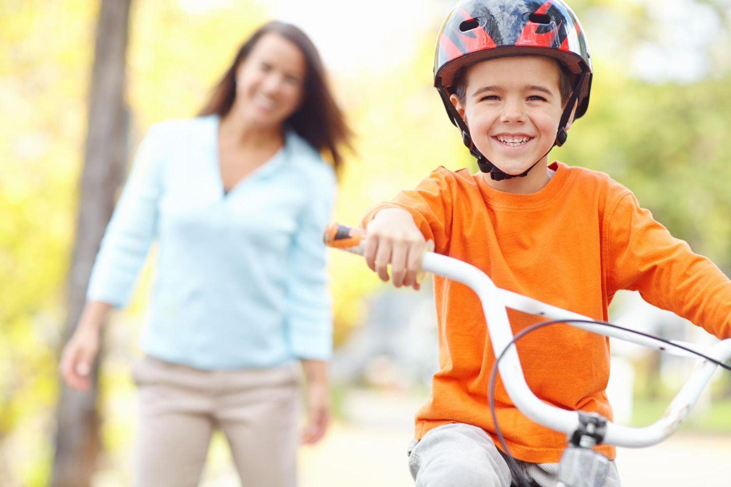 Child in orange helmet and shirt riding a bike, smiling, with an adult walking behind on a sunny path.