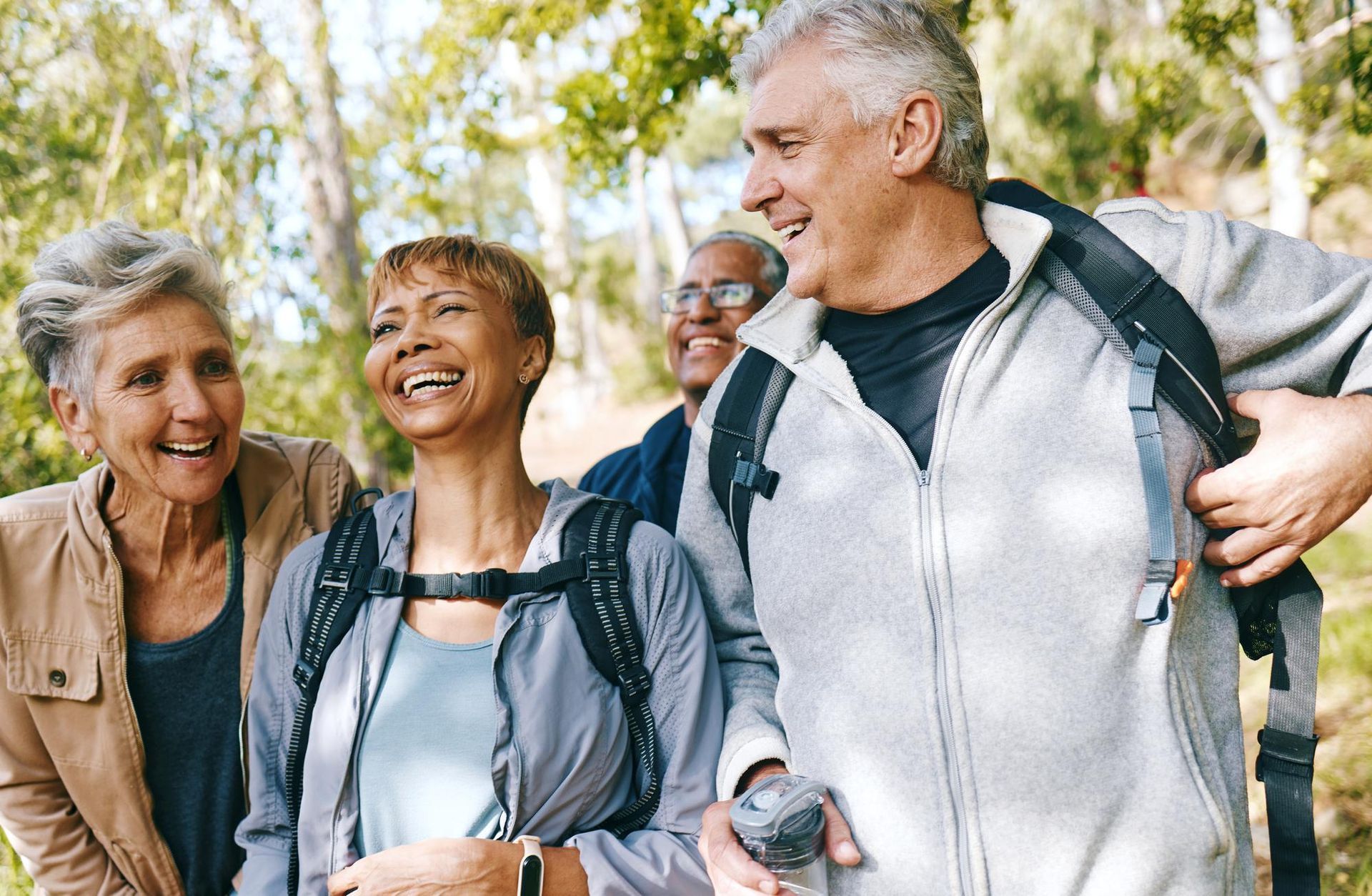 Group of people hiking outdoors, smiling. Trees in the background. Two women and two men wear backpacks.