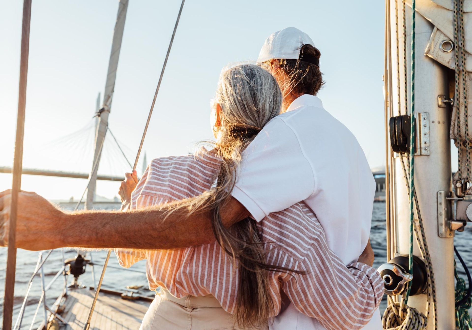 Couple embracing on a sailboat, looking at the water. Sunlight, rigging, and a bridge are visible.