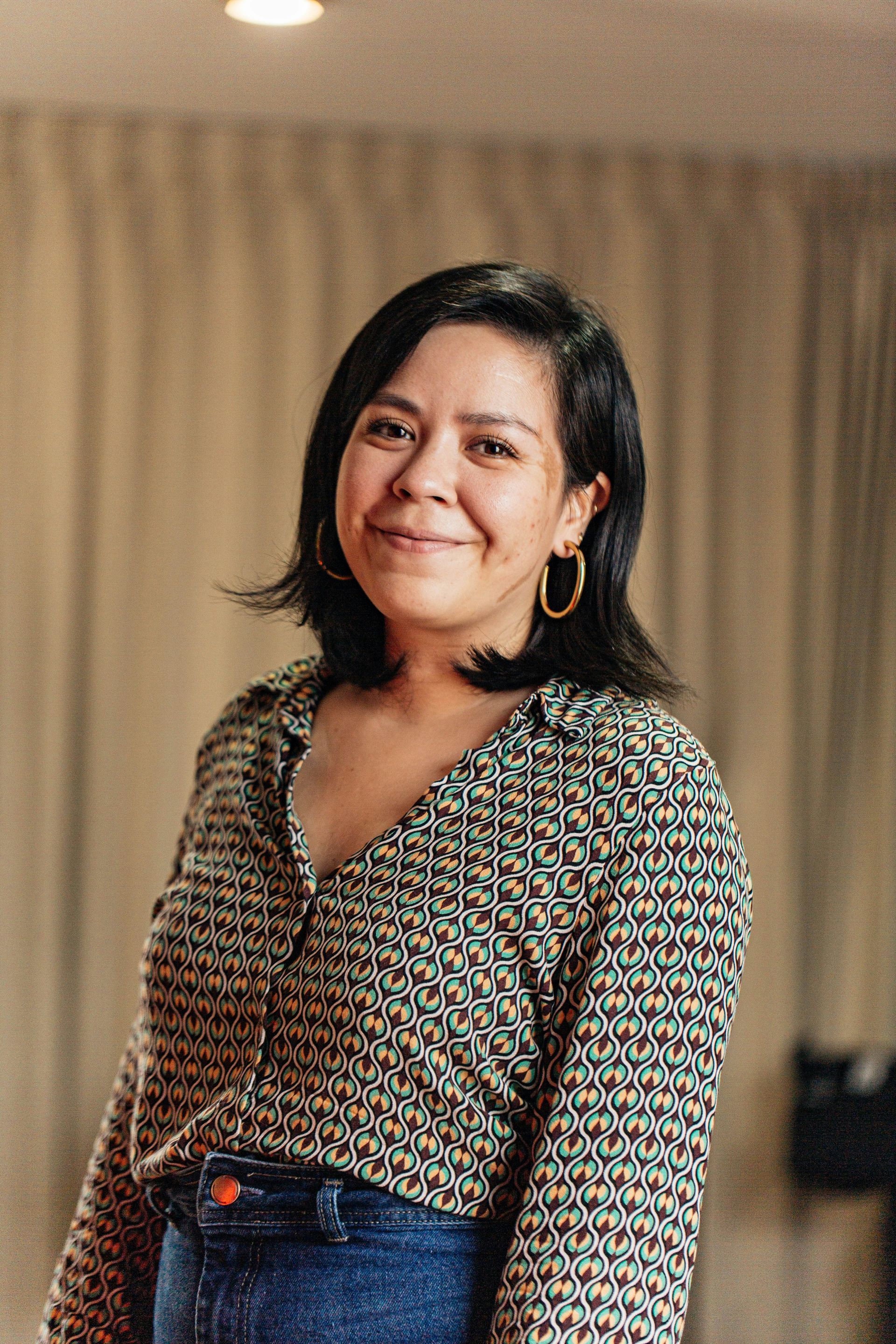 Woman in patterned shirt smiles, standing indoors near curtains.