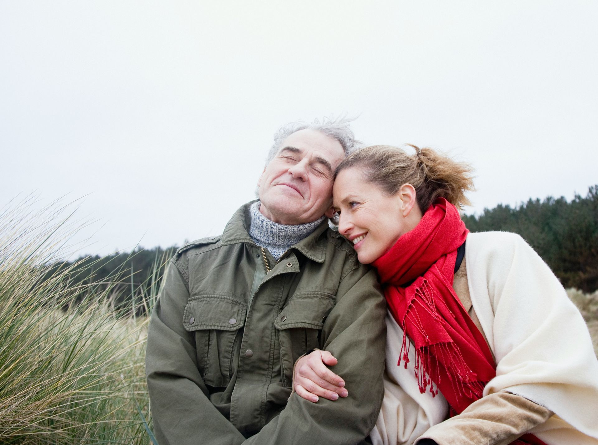 Older couple embraces outdoors; man smiles, woman leans on his shoulder, both warm clothing.