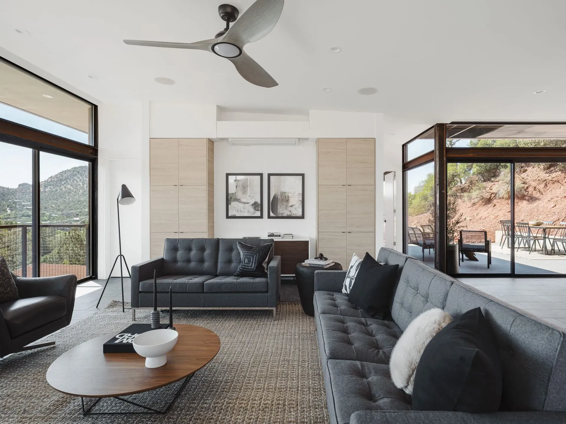 Modern living room with dark sofas, a round coffee table, a ceiling fan, and large windows overlooking a patio.