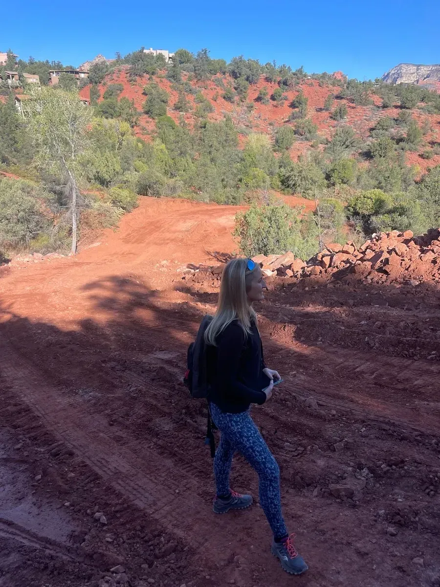 Person standing on a red dirt trail in a rocky, green desert landscape under a clear blue sky