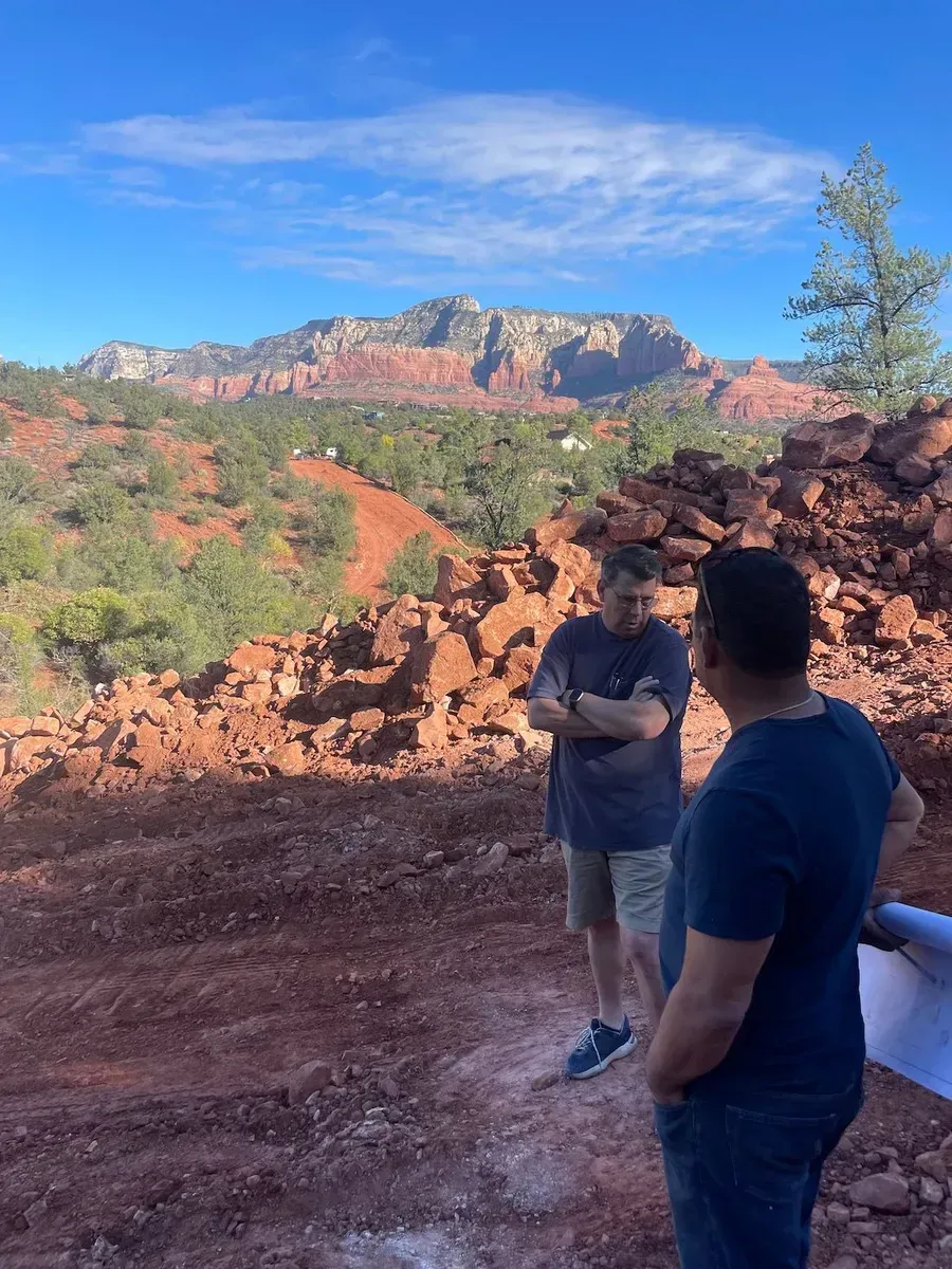 Two people stand on a rocky overlook with red cliffs and blue sky in the background.