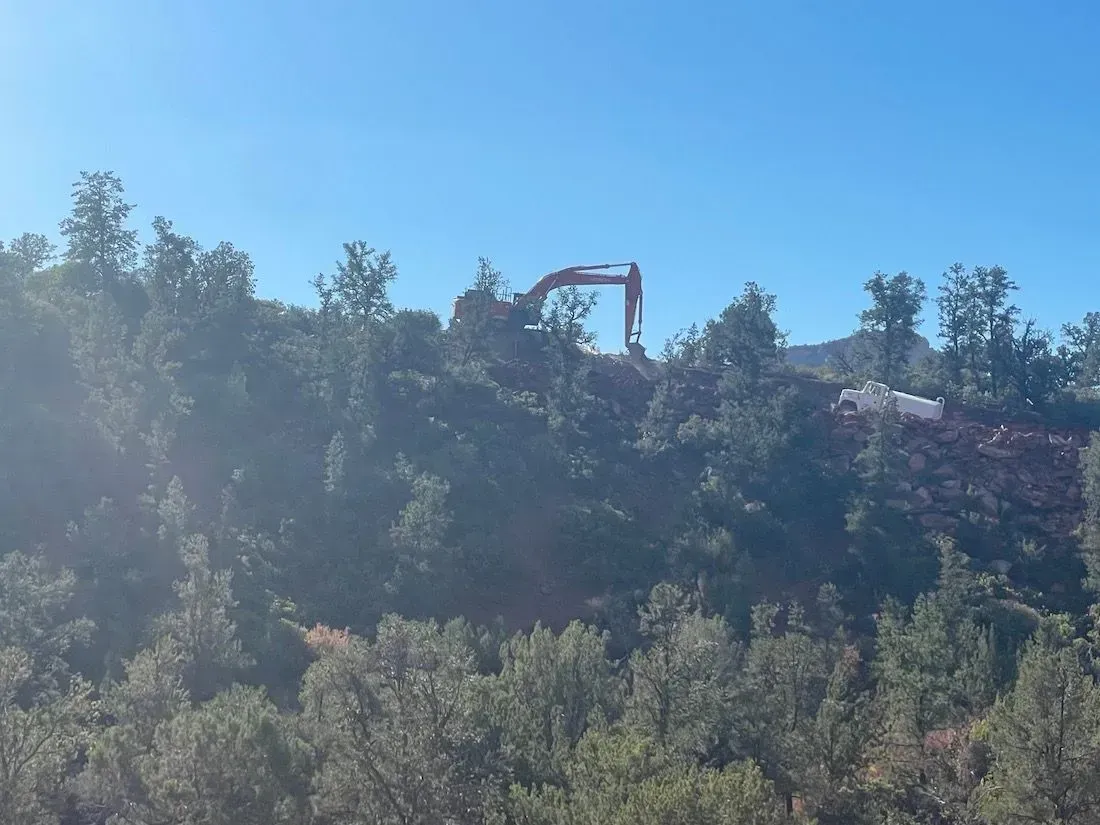 Excavator on a wooded hillside under a clear blue sky, with dust rising from the site