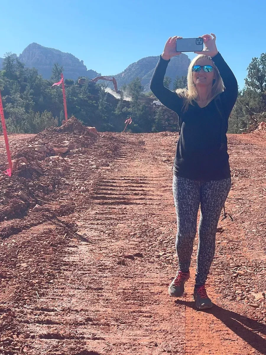 Person in sunglasses taking a selfie on a sunlit dirt path with mountains in the background