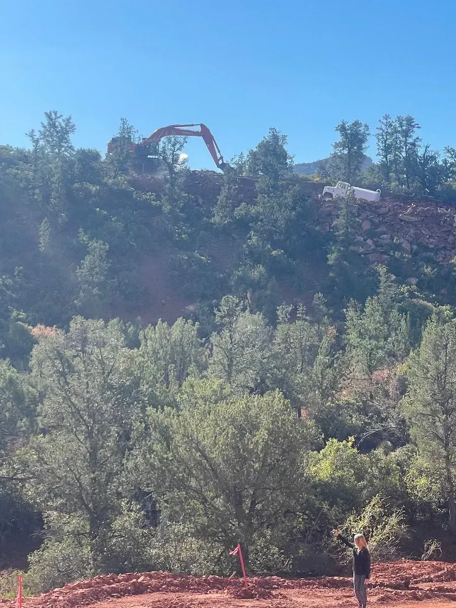 Red rock hillside with trees and a bright blue sky, with two small hikers below