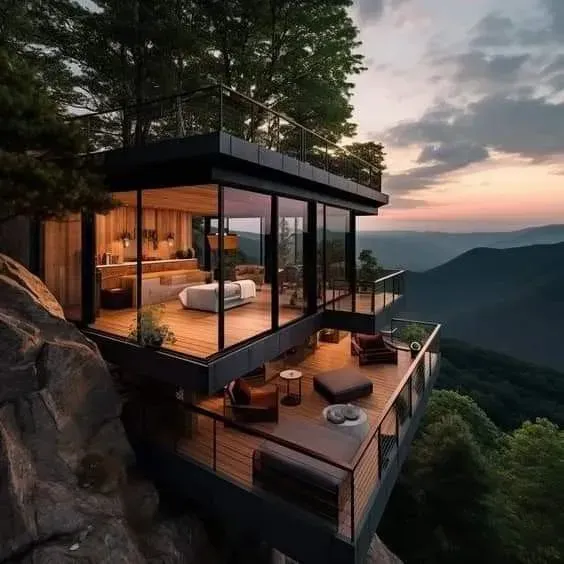 Modern glass mountain villa at sunset with warm interior lights and a balcony overlooking forested valleys