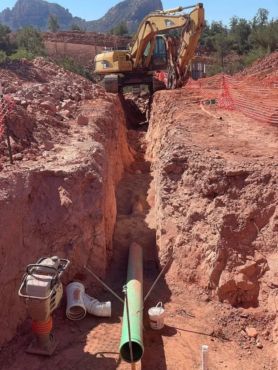 Excavator digging a deep trench with a green pipe laid inside, surrounded by red dirt and orange safety fencing
