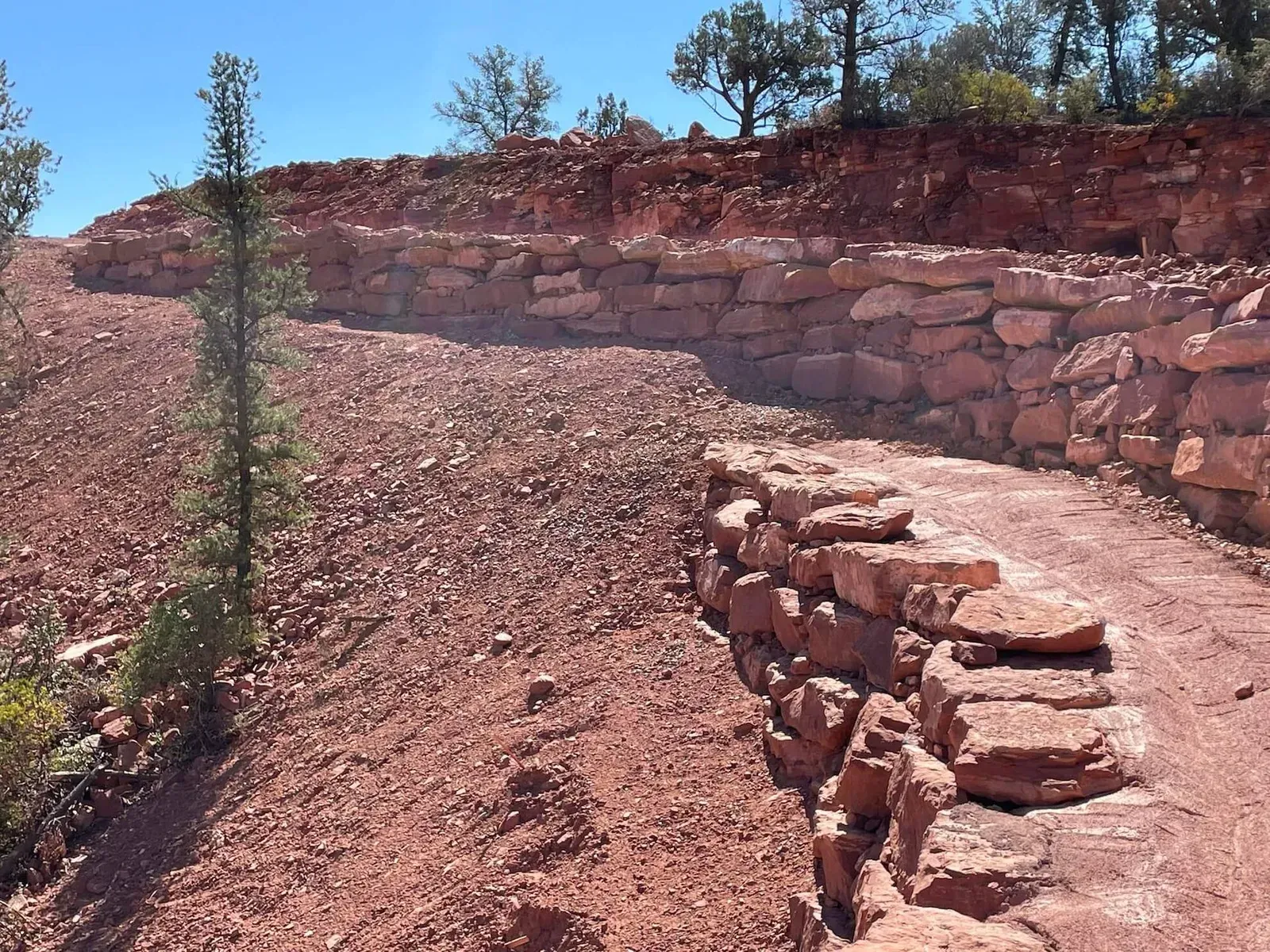 Curved rocky dirt path beside a stone retaining wall under a clear blue sky